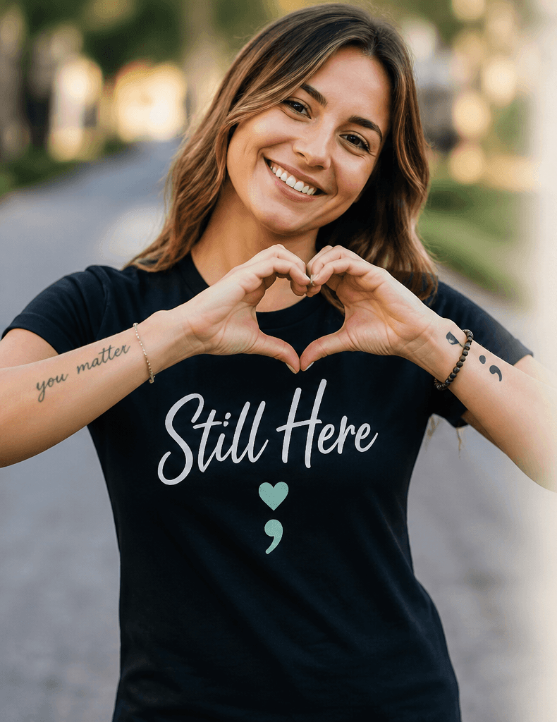 Woman wearing a 'Still Here' shirt with a heart symbol, standing outdoors.